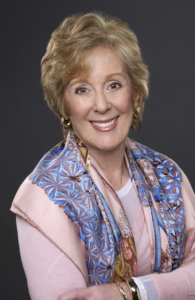 Headshot image of Carol Reichenthal against a gray backdrop. Carol is wearing a pink blouse and a blue draped scarf.